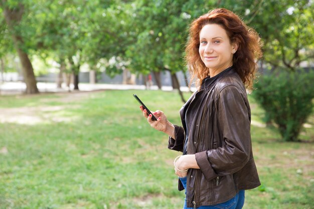 Señora joven positiva que usa el teléfono inteligente en el parque de la ciudad