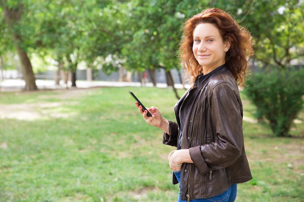 Señora joven positiva que usa el teléfono inteligente en el parque de la ciudad