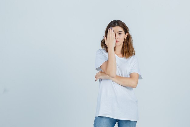 Señora joven que cubre el ojo con la mano en camiseta, jeans y parece serio. vista frontal.