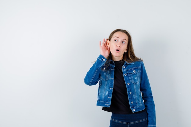 Señora joven que lleva a cabo la mano detrás de la oreja en la blusa y que parece curiosa.