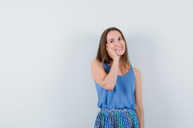 Señora joven que lleva a cabo la mano en la mejilla en camiseta, falda y que parece soñadora. vista frontal.