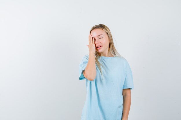 Señora joven que lleva a cabo la mano en el ojo en camiseta y parece linda. vista frontal.