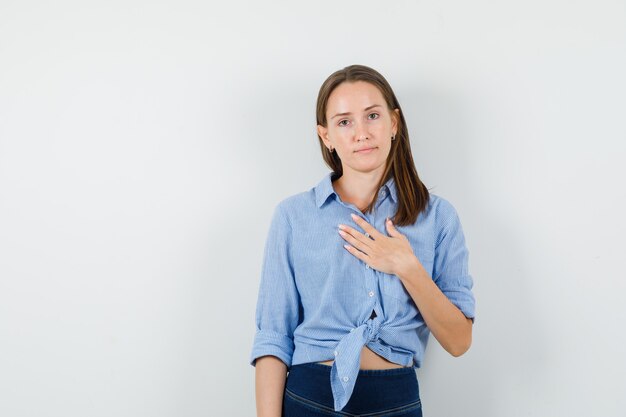 Señora joven que lleva a cabo la mano en el pecho en camisa azul, pantalones y que parece positiva.