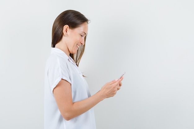 Señora joven que mira su teléfono mientras sonríe en blusa blanca y parece feliz.