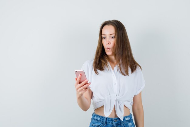 Señora joven que mira el teléfono en la blusa blanca y que parece sorprendida. vista frontal.