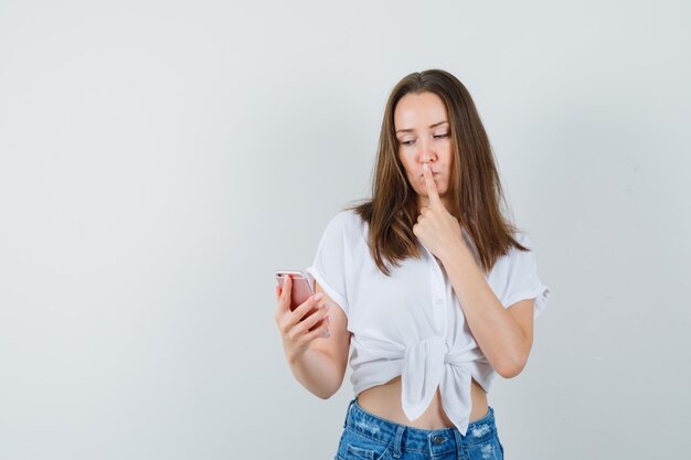 Señora joven que mira el teléfono mientras muestra un gesto silencioso en una blusa blanca y parece tranquilo. vista frontal. espacio para texto