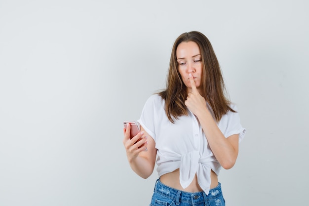 Señora joven que mira el teléfono mientras muestra un gesto silencioso en una blusa blanca y parece tranquilo. vista frontal. espacio para texto