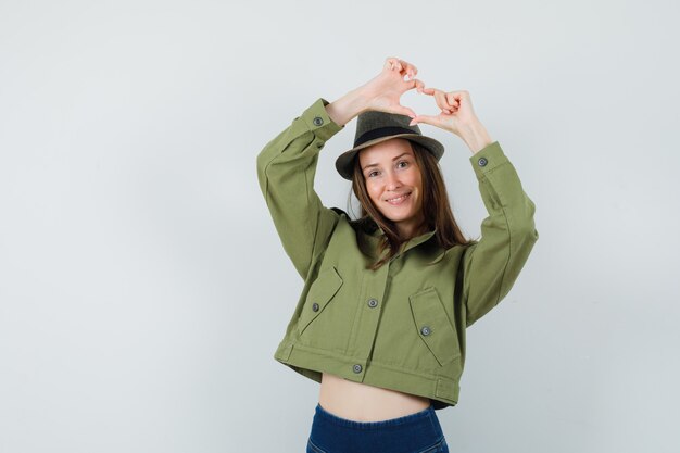 Señora joven que muestra el gesto del corazón en el sombrero de los pantalones de la chaqueta y que mira jovial