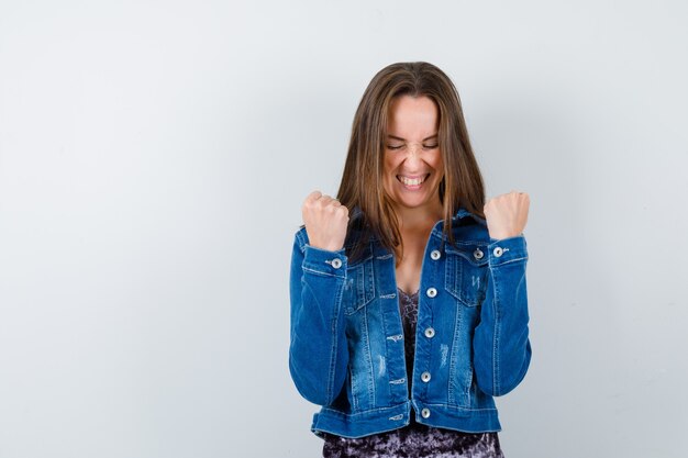 Señora joven que muestra el gesto del ganador en blusa, chaqueta de mezclilla y mirando dichoso, vista frontal.