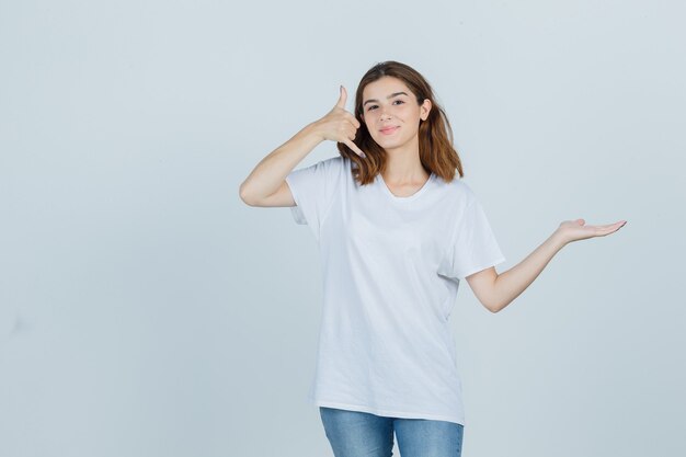 Señora joven que muestra el gesto del teléfono mientras finge sostener algo en una camiseta, jeans y parece confiado, vista frontal.