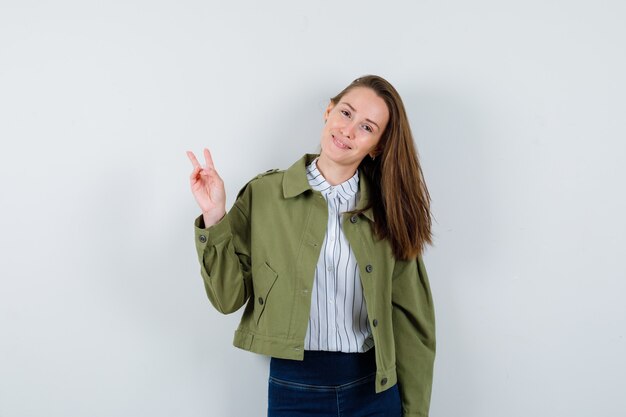 Señora joven que muestra el gesto de la victoria en camisa, chaqueta y que parece confiada. vista frontal.