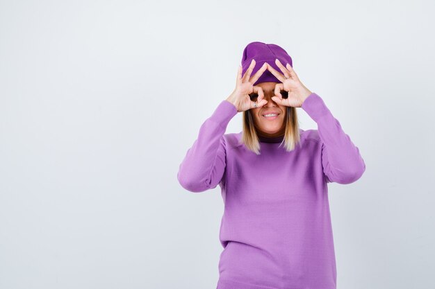 Señora joven que muestra el gesto de los vidrios en suéter púrpura, gorro y que parece divertido. vista frontal.