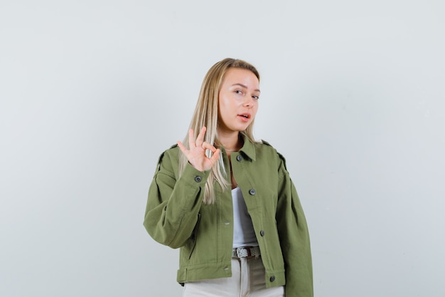 Señora joven que muestra la muestra aceptable en chaqueta, pantalones y parece confiada. vista frontal.