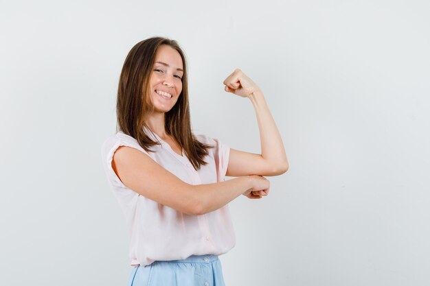 Señora joven que muestra los músculos del brazo en camiseta, falda y parece confiada. vista frontal.