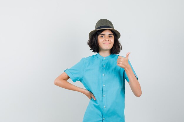 Señora joven que muestra el pulgar hacia arriba en camisa azul, sombrero y parece optimista.