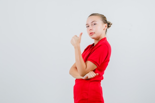 Señora joven que muestra el pulgar hacia arriba en la camiseta roja y parece complacida.