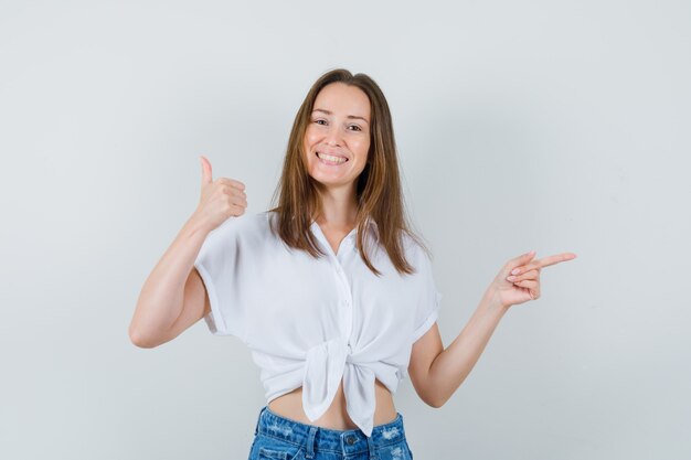 Señora joven que muestra el pulgar hacia arriba mientras apunta a un lado con una blusa blanca y parece optimista. vista frontal.