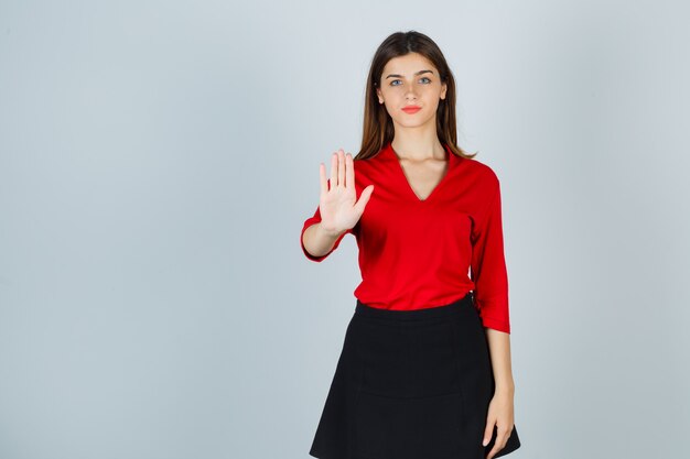 Señora joven que muestra la señal de pare en blusa roja, falda negra y mirando feliz