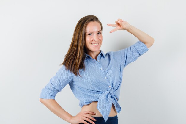 Señora joven que muestra el signo de la victoria en camisa azul, pantalones y aspecto alegre.