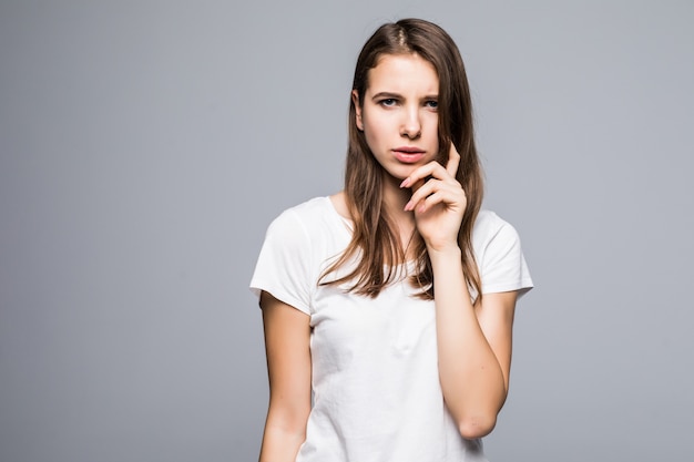 Señora joven que piensa en la camiseta blanca y los pantalones vaqueros azules permanecen delante del fondo blanco del estudio