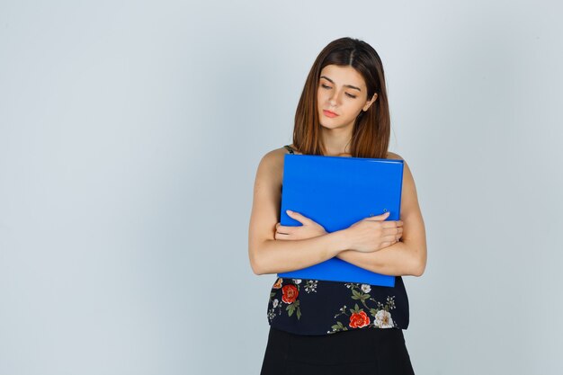 Señora joven que sostiene la carpeta mientras mira hacia abajo en blusa, falda y parece triste. vista frontal.