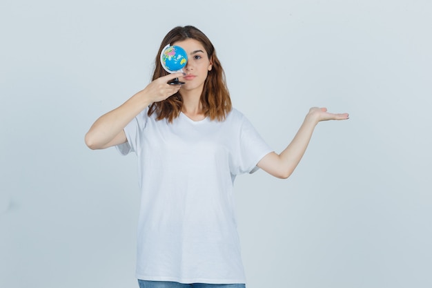 Señora joven que sostiene el globo cerca del ojo mientras muestra algo en camiseta, jeans y parece confiado. vista frontal.