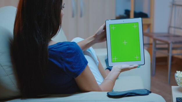 Señora joven que sostiene una tableta con pantalla verde sentado en el sofá. Mujer leyendo, mirando la plantilla chroma key aislado pantalla de portátil inteligente usando tecnología internet acostado en un cómodo sofá.