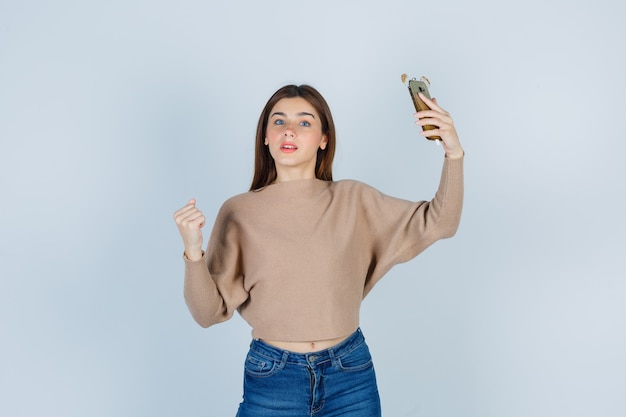 Señora joven que sostiene el teléfono móvil mientras muestra el puño cerrado en un suéter beige, jeans y parece seguro. vista frontal.