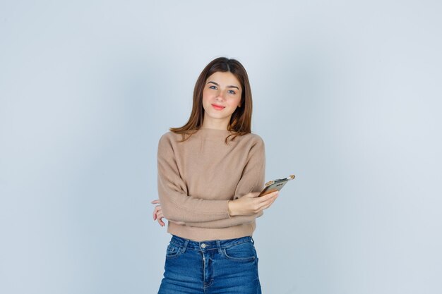 Señora joven que sostiene el teléfono móvil, de pie con los brazos cruzados en un suéter beige, jeans y mirando complacido, vista frontal.
