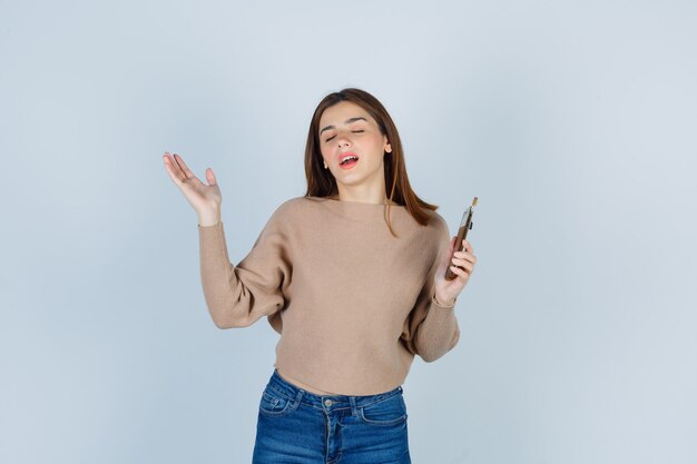 Señora joven que sostiene el teléfono móvil en suéter beige, jeans y parece feliz. vista frontal.