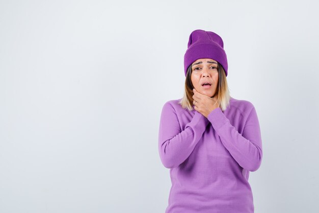 Señora joven que sufre de dolor de garganta en un suéter morado, gorro y con aspecto doloroso. vista frontal.