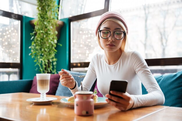 Señora joven que usa el teléfono que bebe el café mientras que se sienta en café.