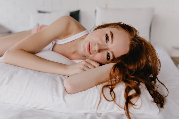 Señora joven romántica con el pelo de jengibre brillante posando sobre la almohada. Foto interior de modelo femenino extático en ropa de dormir blanca.