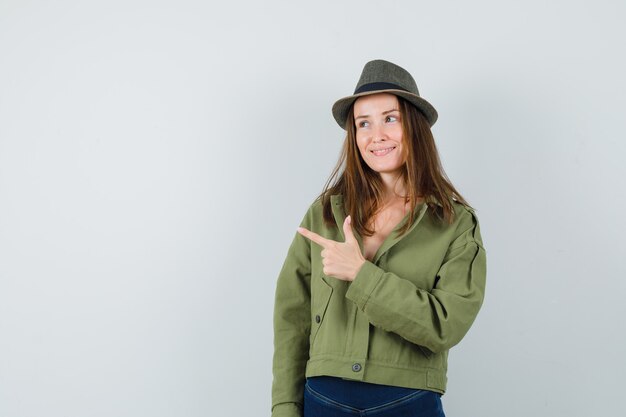 Señora joven con sombrero de pantalones de chaqueta apuntando a la esquina superior izquierda y mirando soñadora