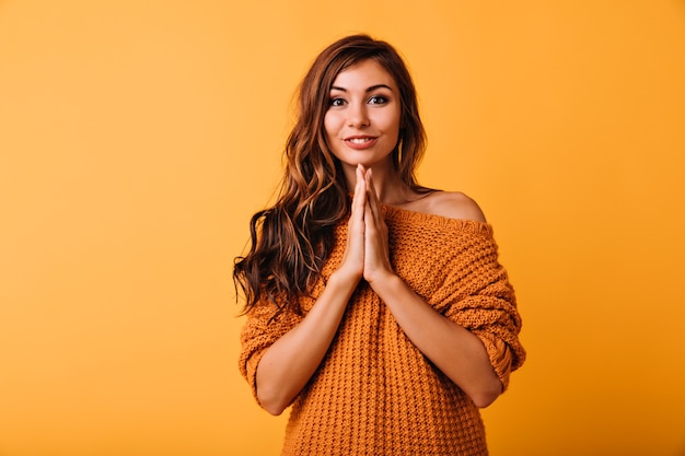 Señora joven soñadora en elegante suéter naranja posando con linda sonrisa. Retrato de interior de adorable niña caucásica con largo cabello ondulado.