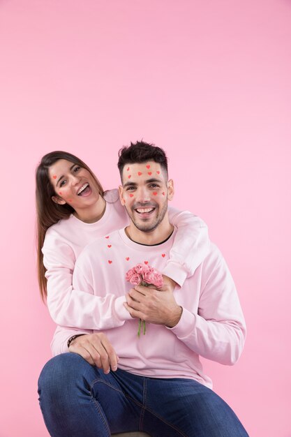 Señora joven sonriente con las flores que abrazan al individuo feliz con los corazones de papel en cara