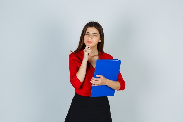 Señora joven sosteniendo la carpeta mientras está de pie en pose de pensamiento en blusa roja