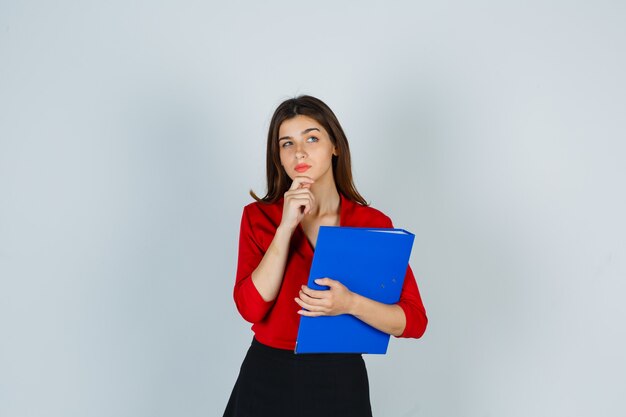 Señora joven sosteniendo la carpeta mientras está de pie en pose de pensamiento en blusa roja