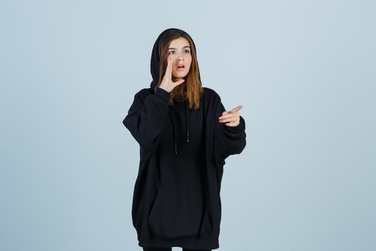 Foto gratuito señora joven con sudadera con capucha de gran tamaño, pantalones apuntando a un lado mientras grita algo con la mano y mira perpleja, vista frontal.