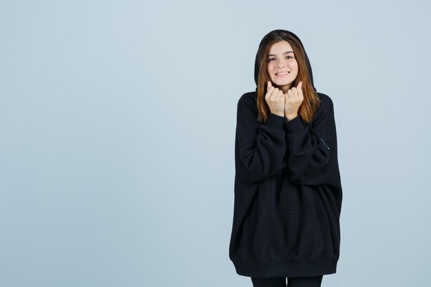 Señora joven con sudadera con capucha de gran tamaño, pantalones sosteniendo los puños cerca de la cara y luciendo linda vista frontal.