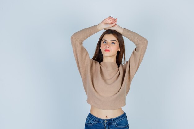 Señora joven en suéter beige, jeans con las manos sobre la cabeza y mirando confiado, vista frontal.