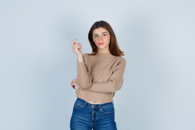 Señora joven en suéter beige, jeans posando mientras mira al frente y mirando confiada, vista frontal.