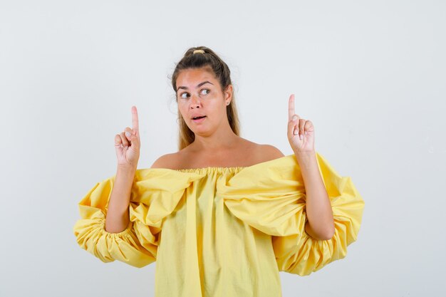 Señora joven en vestido amarillo apuntando hacia arriba y mirando pensativo, vista frontal.