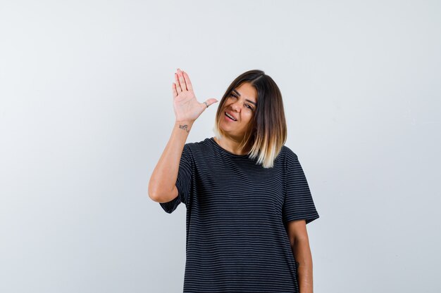 Señora joven en vestido de polo que muestra la palma de la mano para saludar y mirar alegre, vista frontal.