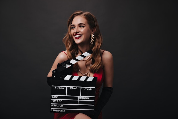 Señora con labios rojos sosteniendo claqueta sobre fondo negro Mujer feliz en vestido de seda de lujo sonriendo y posando sobre fondo aislado