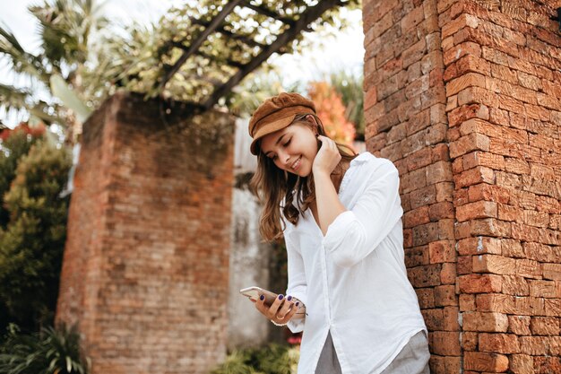 Señora con manicura azul con smartphone. Chica de blusa blanca y pantalón gris posando junto a la pared de ladrillo con plantas tropicales.