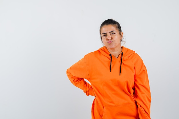 Señora manteniendo la mano en la cintura, labios curvados, cara con el ceño fruncido en una sudadera con capucha naranja y luciendo vacilante