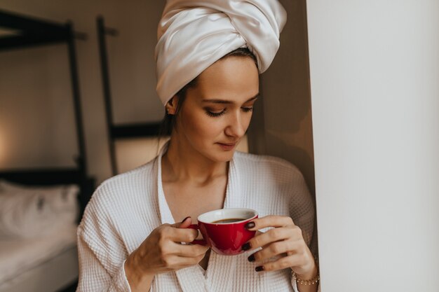 Señora sin maquillaje con una toalla en la cabeza sosteniendo una taza de café roja. Mujer en bata de baño posando en el dormitorio.