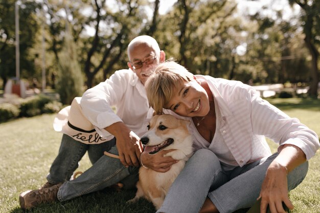 Señora maravillosa con peinado rubio fresco en blusa a rayas y jeans sonriendo y posando con perro y esposo en camisa blanca en el parque.