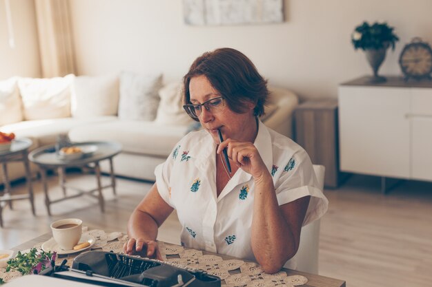 Señora mayor en camisa blanca mordiendo pluma y pensando en casa durante el día